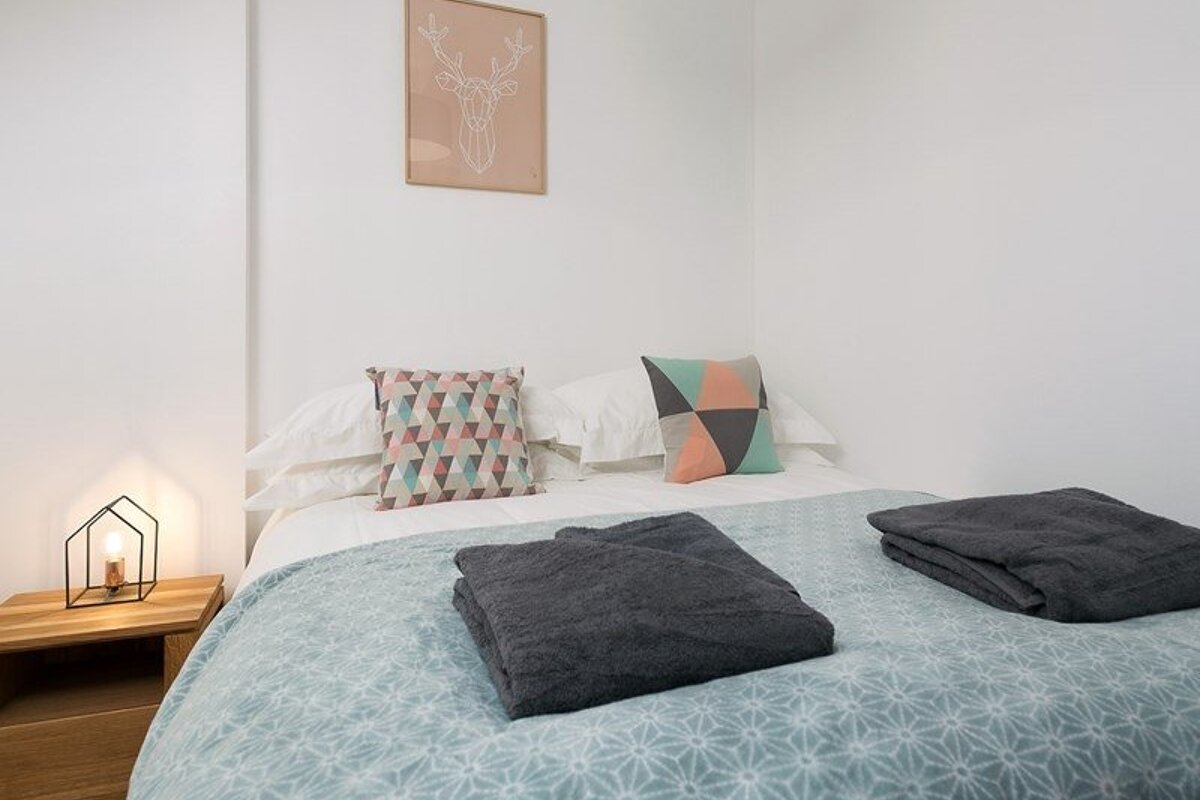 A cozy, modern bedroom featuring a bed with geometric pillows, folded grey towels, a bedside lamp, and a minimalist deer print on the wall.