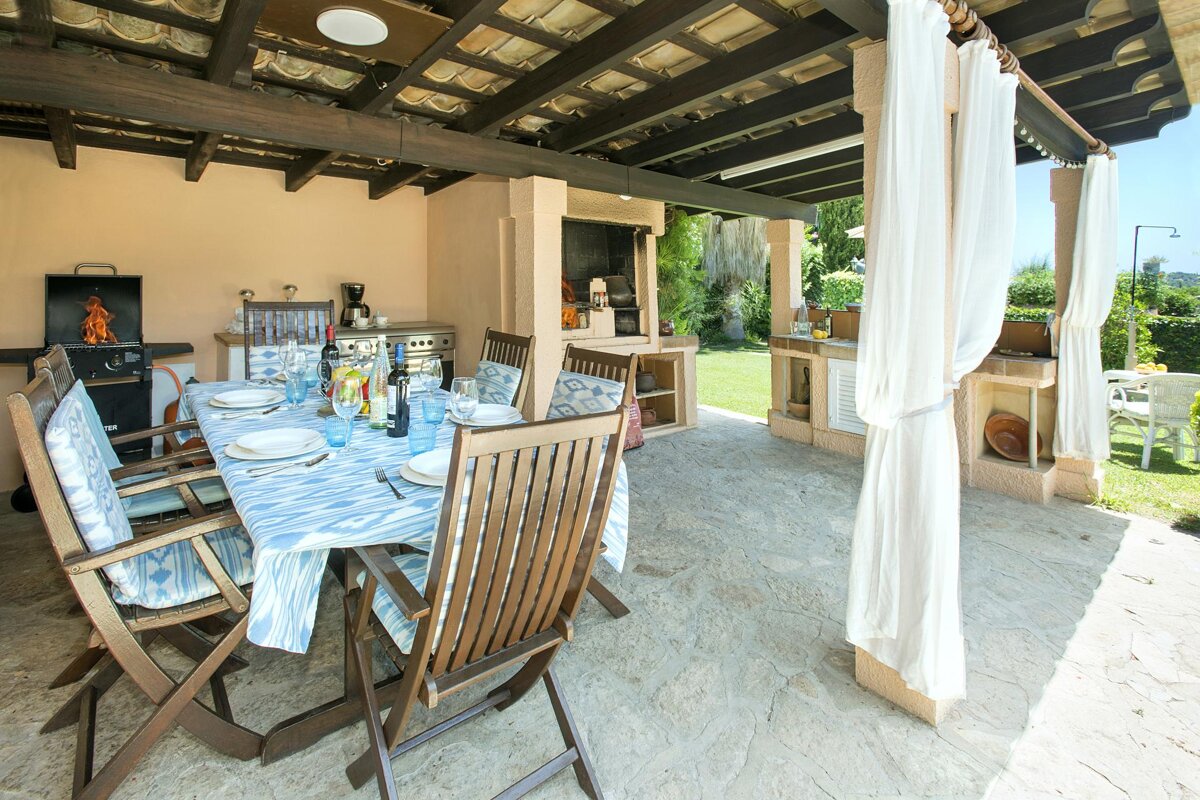 A spacious, covered outdoor patio features a dining table set for guests, multiple BBQ grills (one with active flames), and sheer white curtains, all overlooking a lush garden.