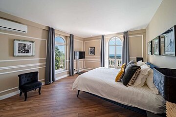 Elegant bedroom featuring a large bed, arched windows/balcony overlooking a landscape, wood floors, and tan walls with decorative trim.