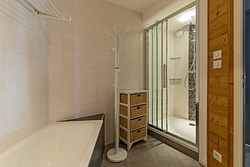 A modern bathroom features a bathtub, glass shower, white tiled walls, grey floor, a white stand, wicker drawers, and wooden paneling.