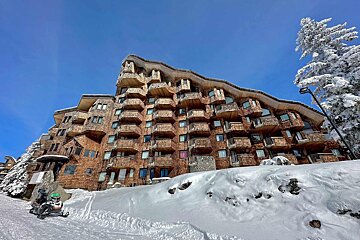 A large wooden apartment building with numerous balconies and ground-floor garages, set against a backdrop of green pine trees.