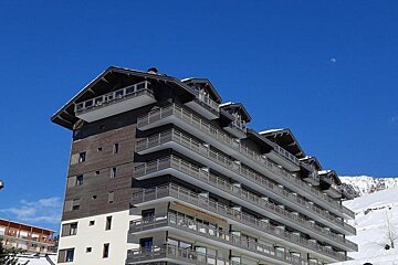 A multi-story wooden building with balconies, a row of garages below, and forest trees in the background.