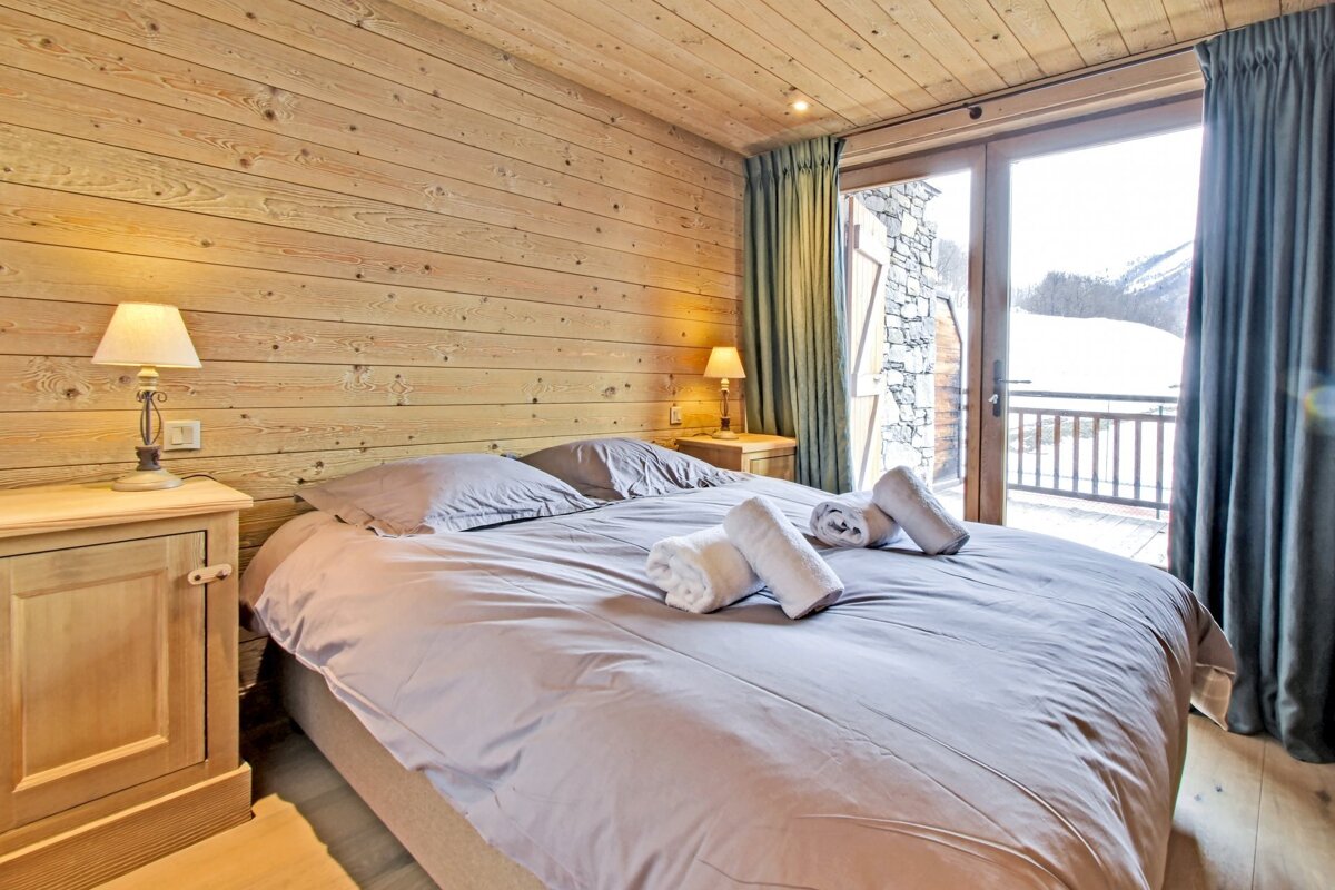 Cozy wooden chalet bedroom featuring a large bed, bedside lamps, and glass doors opening to a snowy winter view.