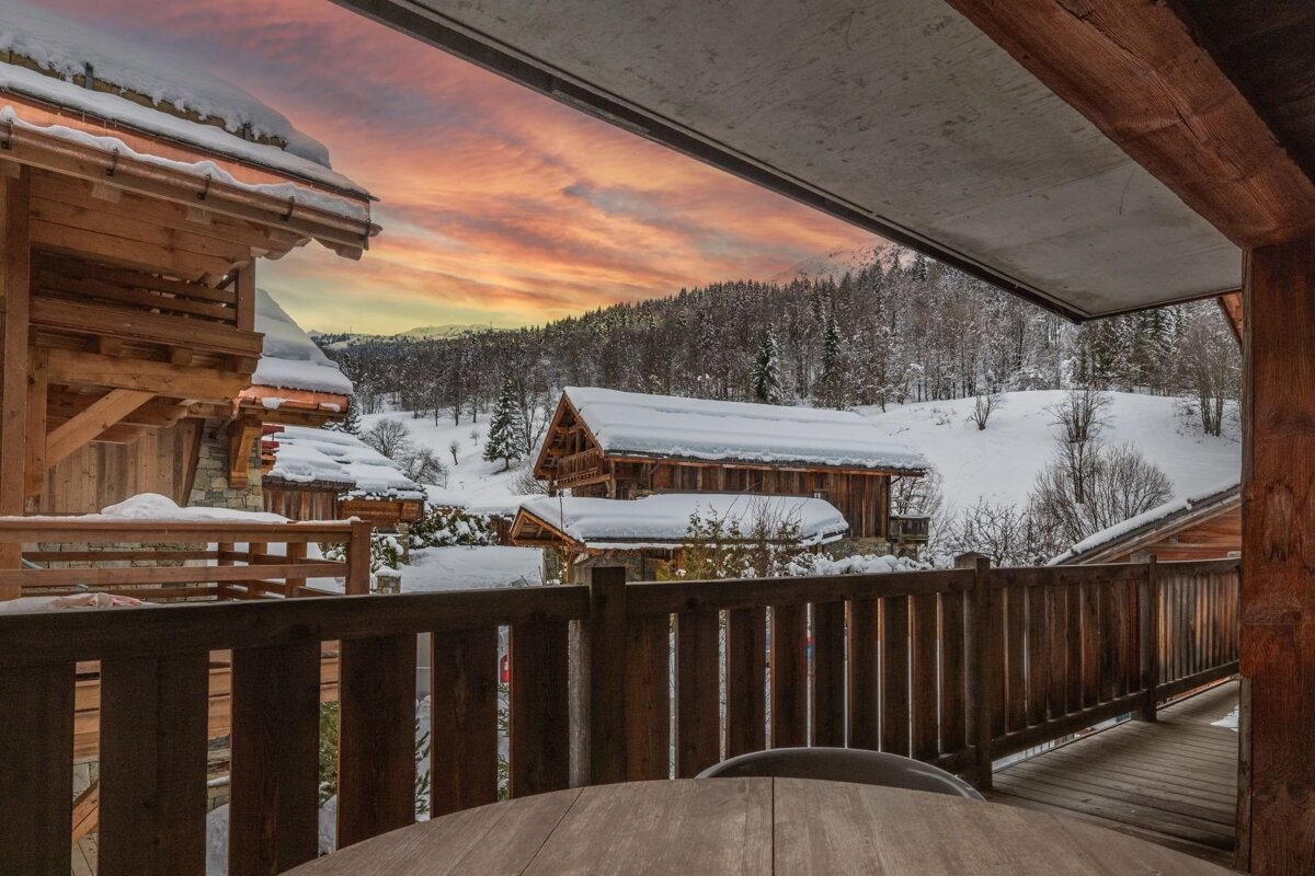View from a balcony showing snow-covered wooden chalets and a forest under a vibrant orange and yellow sunset sky in a winter landscape.