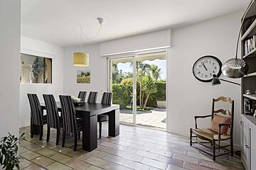 A bright dining room with a black table, striped chairs, and tiled floor. Large glass doors open to a sunny garden. Features a wall clock and old wooden chair.