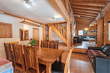 Cozy wooden cabin interior featuring a dining table and chairs, a staircase, and a living room with leather sofas in the background.
