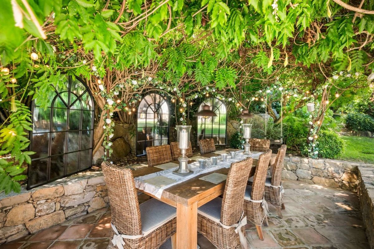 Lush outdoor dining patio with a wooden table, wicker chairs, and arched mirrors, all under a leafy canopy strung with lights and white flowers.