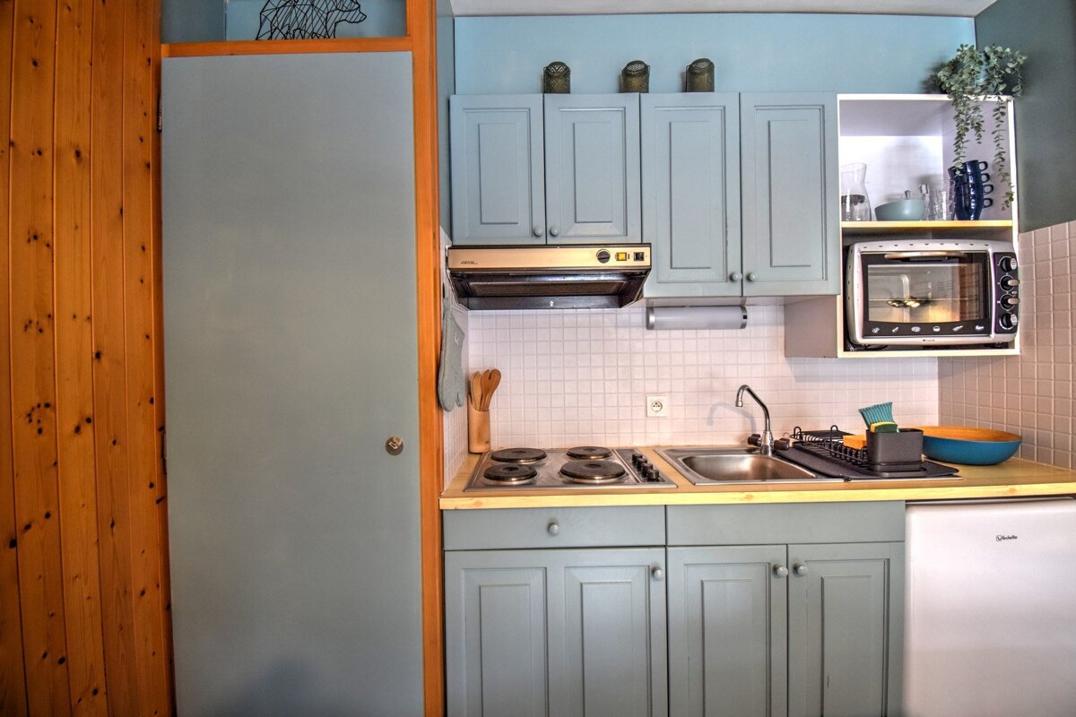 A cozy, compact kitchen with light blue cabinets, a white tiled backsplash, stove, sink, microwave, and mini-fridge. Wood paneling lines the left wall.