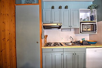A cozy, compact kitchen with light blue cabinets, a white tiled backsplash, stove, sink, microwave, and mini-fridge. Wood paneling lines the left wall.