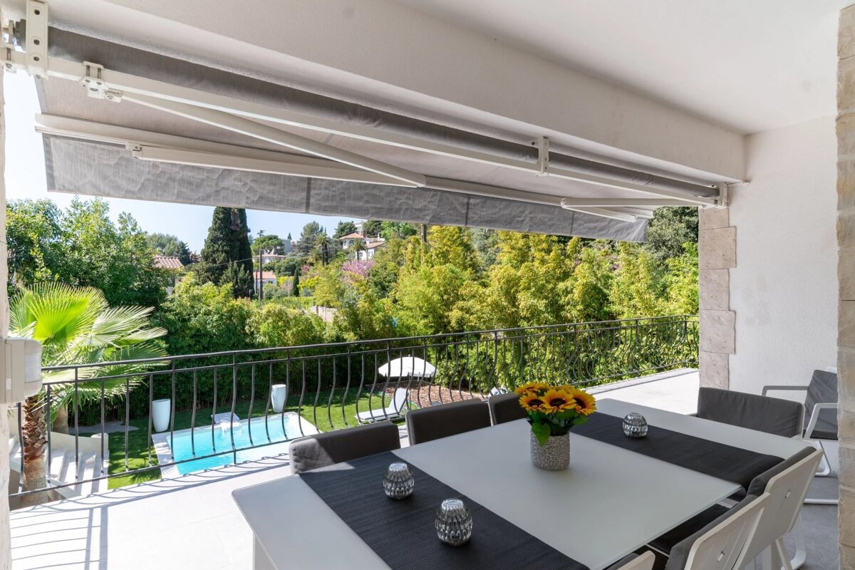 A covered balcony dining area overlooks a lush green garden with a small swimming pool below.