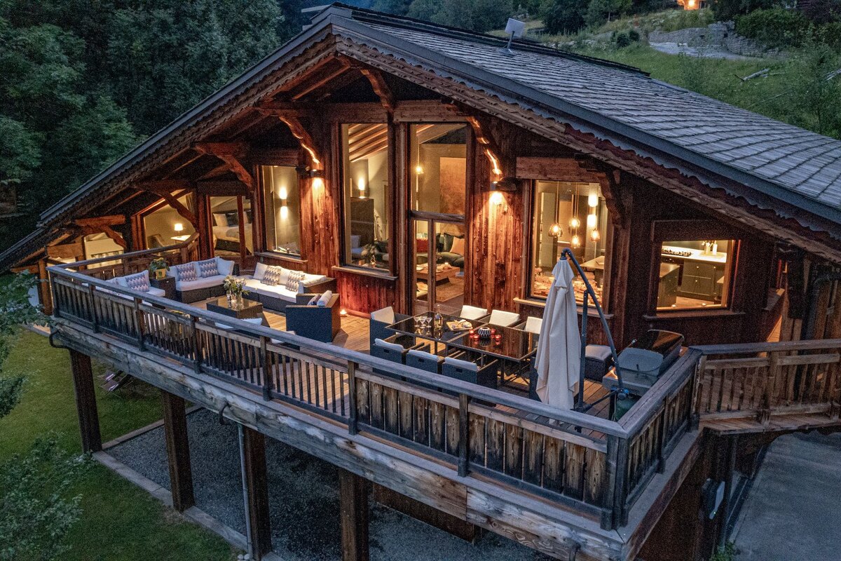 A cozy wooden chalet at dusk, with a large illuminated deck featuring outdoor seating and dining areas, nestled in a wooded setting.