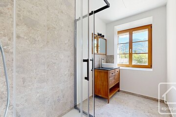 A bright, modern bathroom with a glass shower, wooden vanity and sink, and a window overlooking mountains. Tiled walls and floor in light stone.