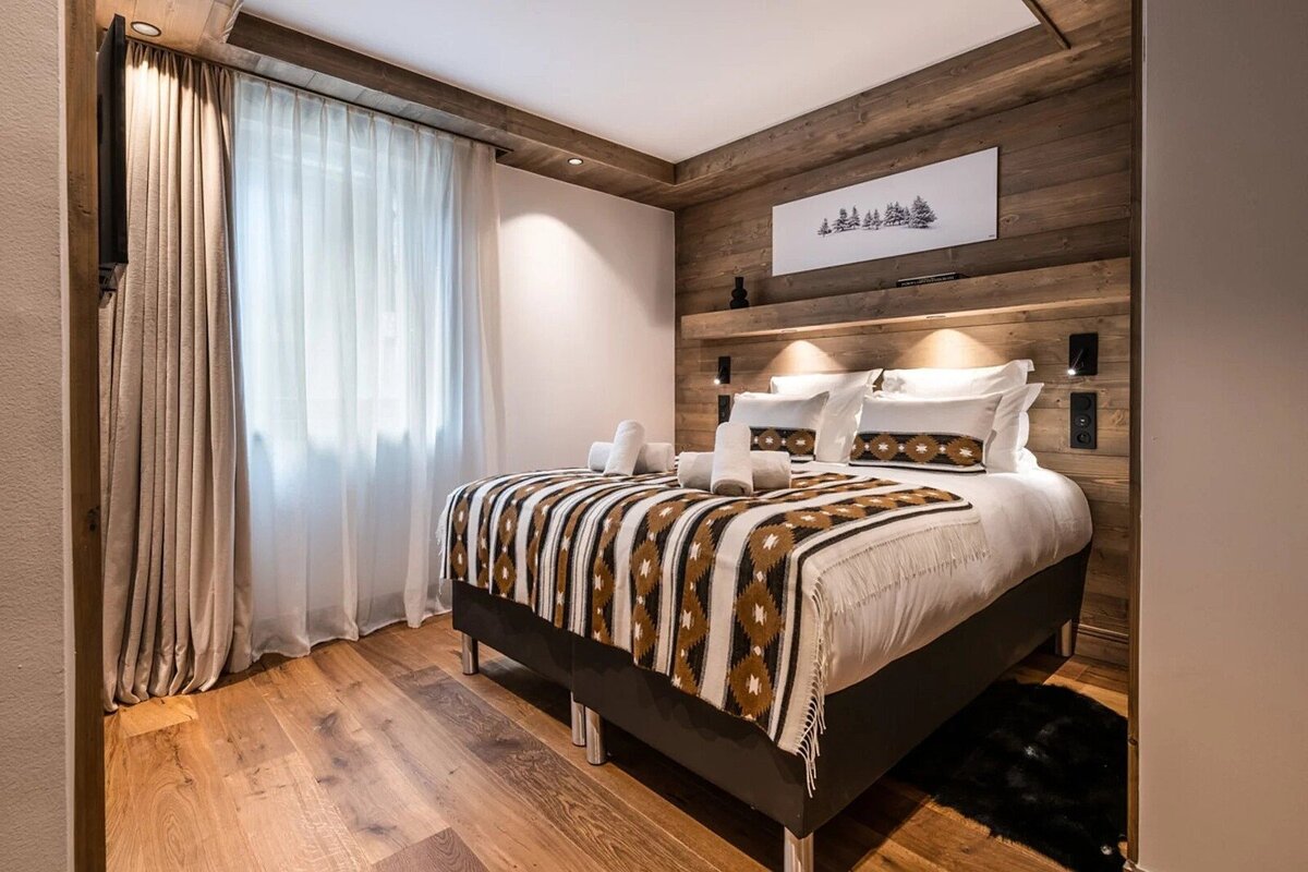 A cozy, modern chalet bedroom with wood paneling, a bed with a geometric patterned blanket, hardwood floors, and a bright window.