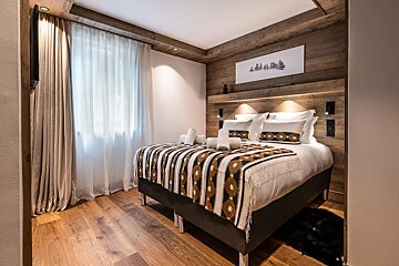 A cozy, modern chalet bedroom with wood paneling, a bed with a geometric patterned blanket, hardwood floors, and a bright window.