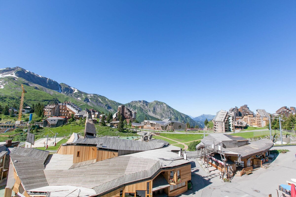 Sunny mountain village with wooden buildings, green hills, distant snow-capped mountains, and a clear blue sky.