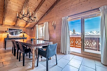 Alpine chalet dining room with wooden walls, antler chandelier, heavy table, and a balcony overlooking snowy mountains.