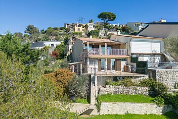 A multi-level villa with balconies on a lush green hillside, surrounded by trees and other houses under a clear blue sky.
