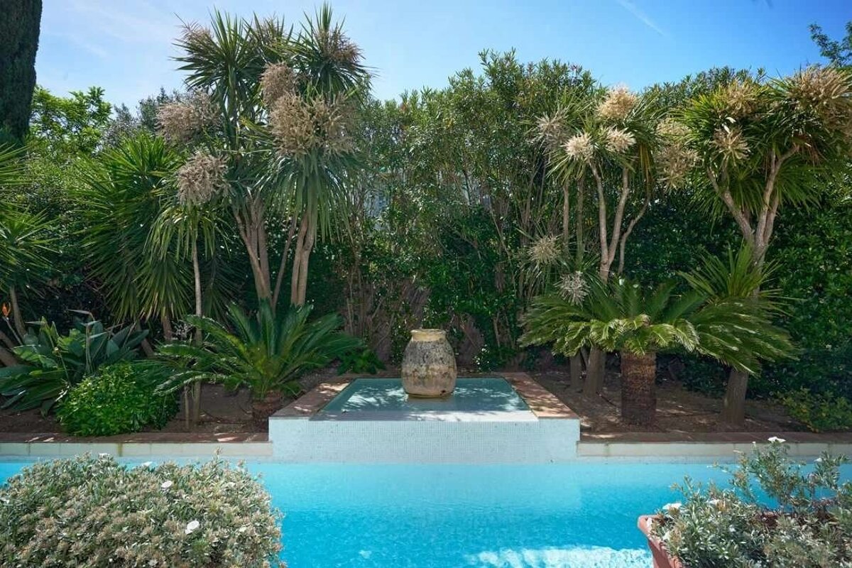 A bright blue pool with a water feature and vase, surrounded by lush tropical palm trees and green plants under a clear sky.