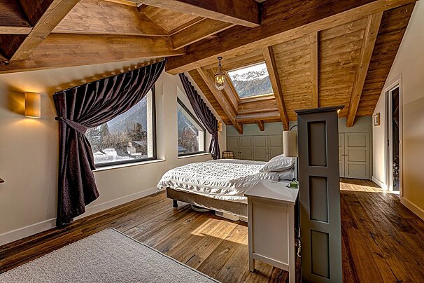 Cozy chalet bedroom with high wood-beam ceilings, a skylight, and large windows offering scenic views of snowy mountains.