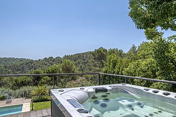 An outdoor hot tub on a deck overlooking dense green forests and rolling hills under a clear blue sky.