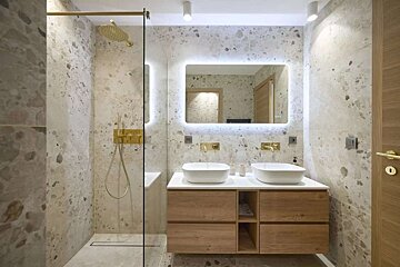 Modern bathroom with speckled walls, gold fixtures, a glass shower, and a double wooden vanity under an illuminated mirror.