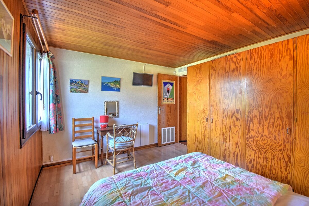 Cozy bedroom featuring warm wood paneling on ceiling/walls, colorful quilt, large wardrobe, window, small desk with chairs & art.