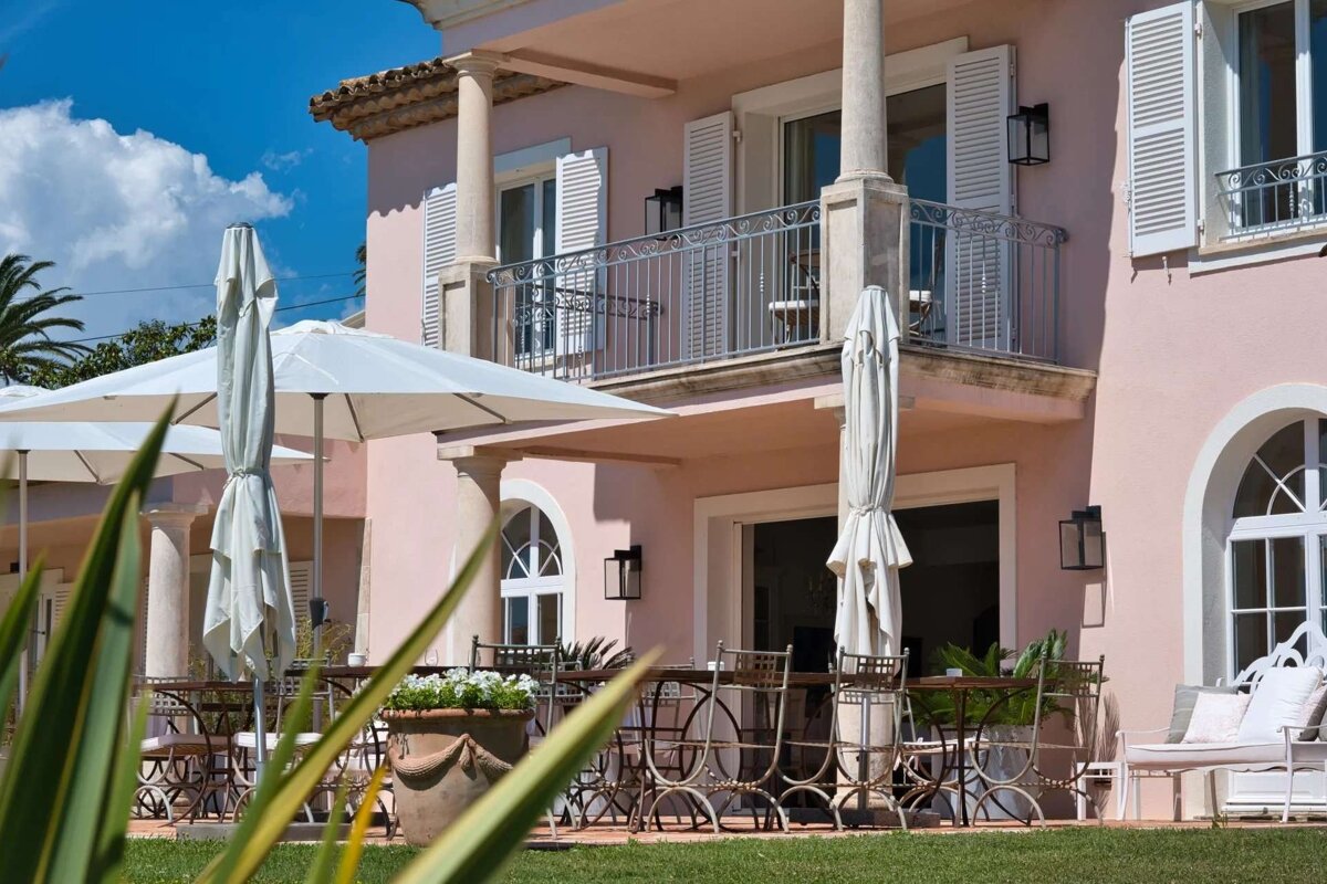 A pink building with an outdoor dining area, white umbrellas, tables, and chairs, featuring a balcony under a bright blue sky.