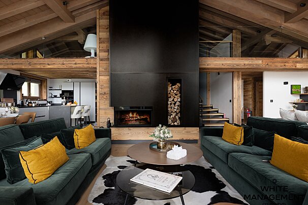 Cozy modern chalet interior with dark green sofas, yellow pillows, a cowhide rug, and a tall black central fireplace.