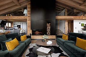 Cozy modern chalet interior with dark green sofas, yellow pillows, a cowhide rug, and a tall black central fireplace.