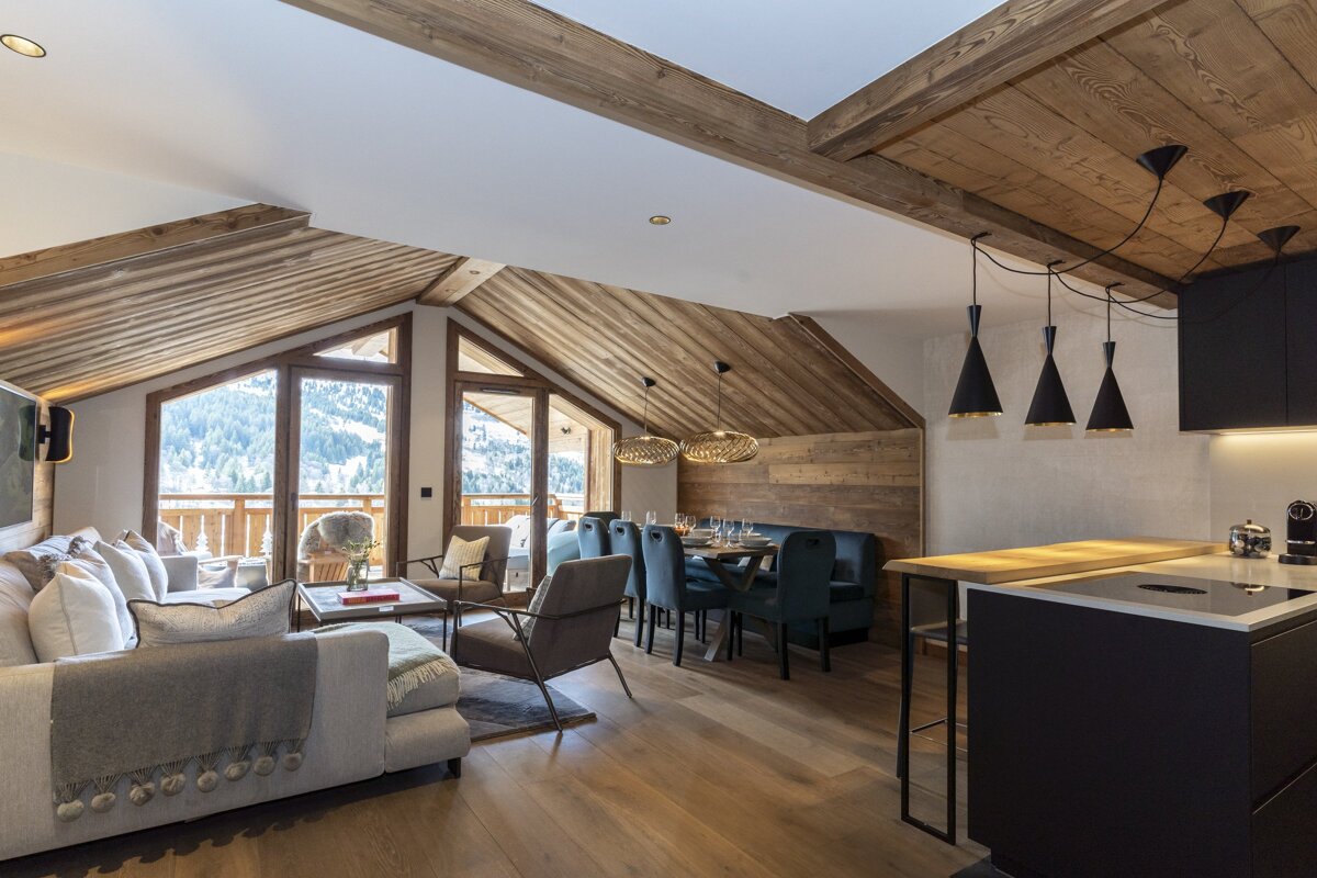 Modern alpine chalet with open living, dining, and kitchen. Vaulted wood ceilings, large windows overlooking snowy mountains, and contemporary decor.