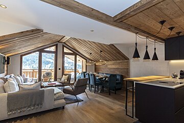 Modern chalet with rustic wood interior, open living/dining/kitchen, and large windows revealing a snowy mountain view.