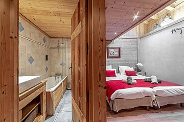 A rustic chalet room with a bathroom (tub, tiled walls) on the left, and a bedroom (twin beds with red accents, wood paneling) on the right, separated by a wooden partition.