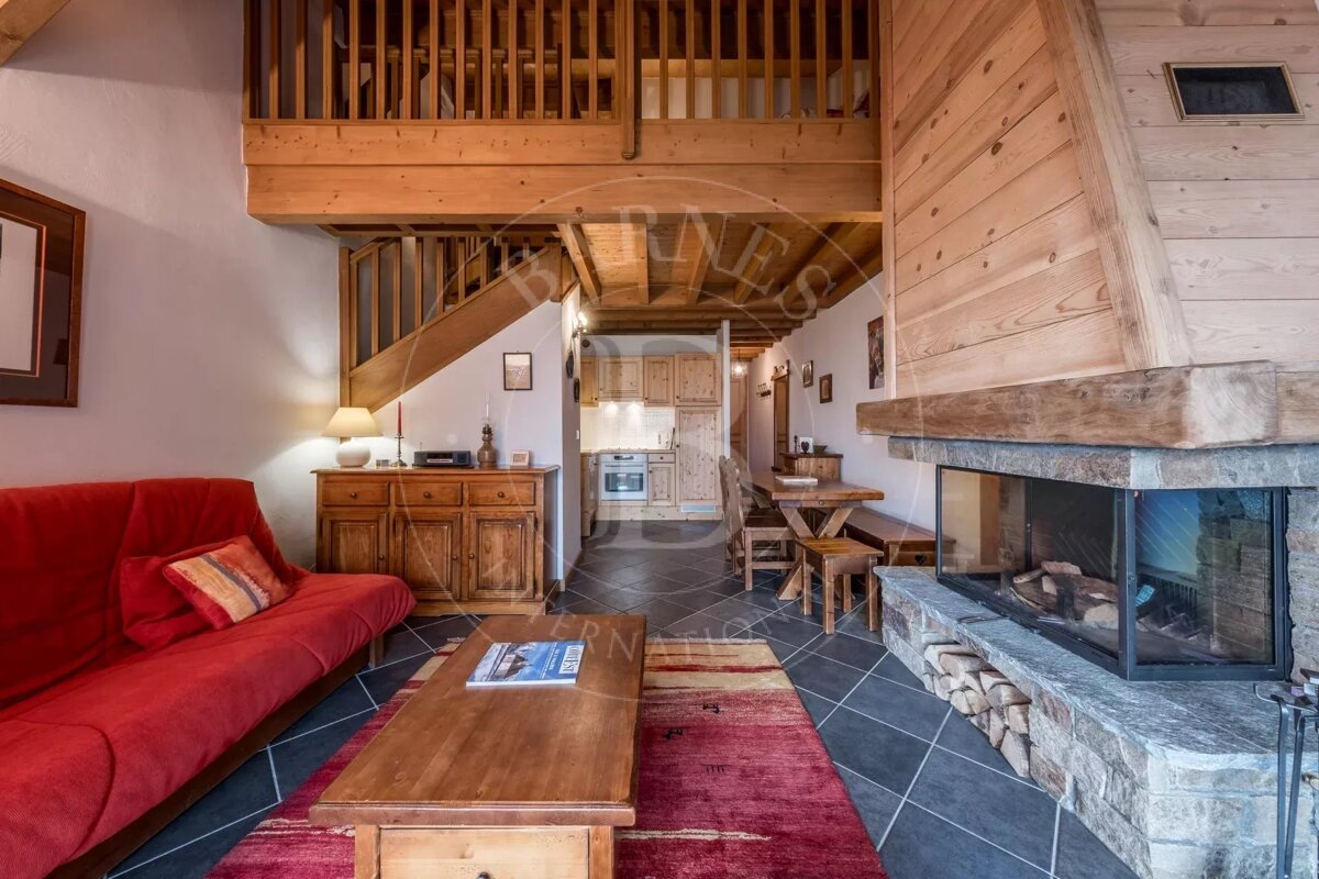 A cozy, rustic chalet living room with a red sofa, large stone fireplace, wooden mezzanine, and an open-plan kitchen/dining area.
