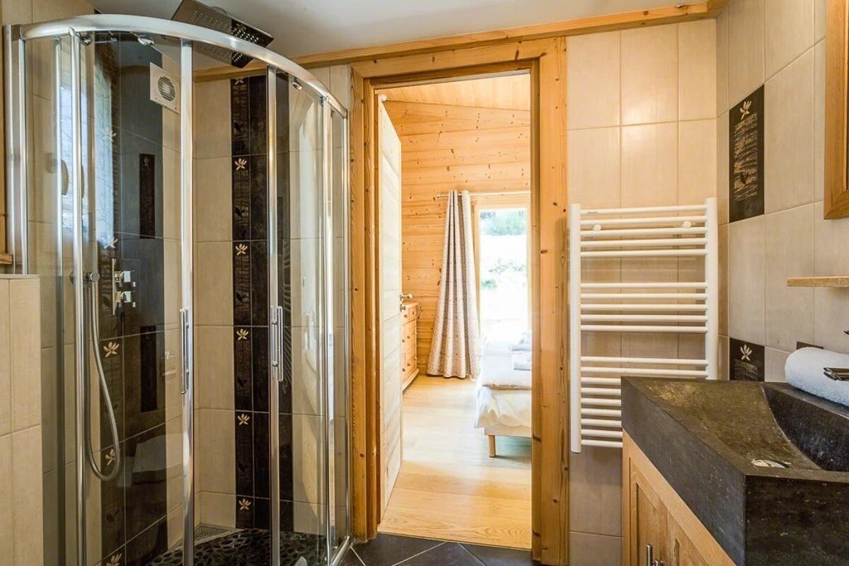 A wooden bathroom with a glass shower enclosure and a view of an adjacent bedroom.
