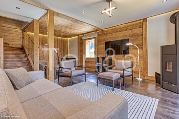 A modern chalet living room with extensive wood paneling, a grey sectional sofa, armchairs, a fireplace, and a visible staircase.
