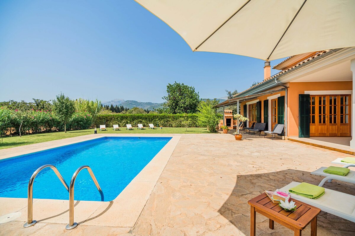 Sunny villa with a blue pool, stone patio, green lawn, and distant mountains. An umbrella shades lounge chairs on a bright day.