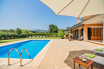 Sunny villa with a blue pool, stone patio, green lawn, and distant mountains. An umbrella shades lounge chairs on a bright day.