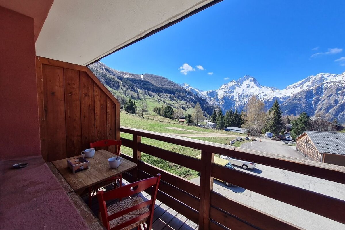 A wooden balcony with a table and red chairs overlooks a stunning green valley and snow-capped mountains under a clear blue sky.