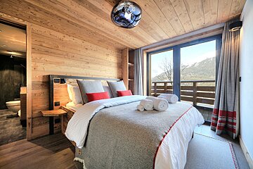 A cozy, modern bedroom with wooden walls and ceiling, a large bed, and an ensuite bathroom, boasting a stunning mountain view.
