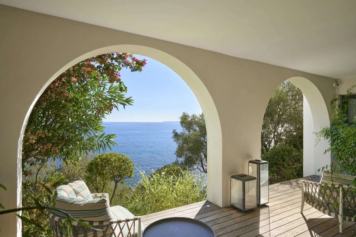 A sunny arched patio offers stunning ocean views, lush greenery, and outdoor seating on a wooden deck, creating a serene Mediterranean scene.