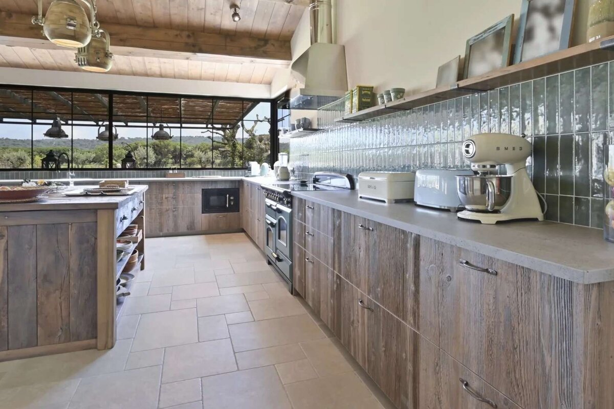A spacious rustic-chic kitchen with weathered wood cabinets and a large island. Panoramic windows reveal a lush outdoor view, complemented by light green subway tiles and modern appliances.
