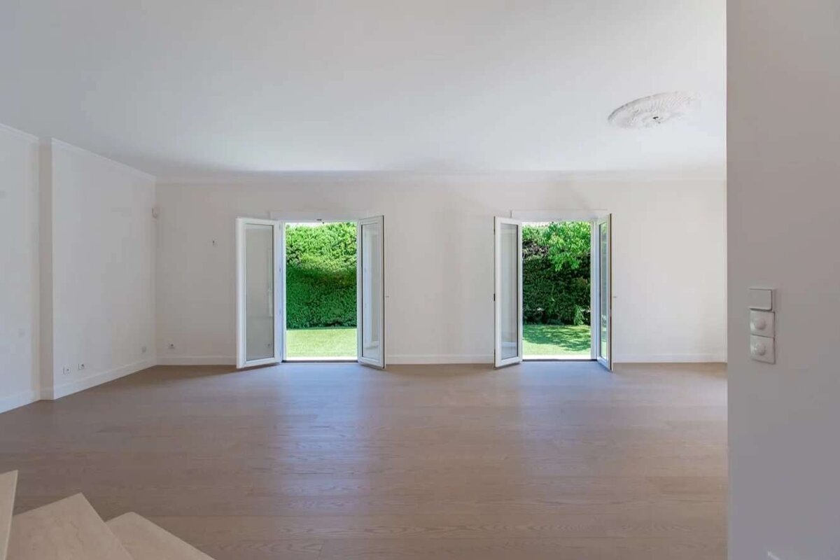 An empty, bright room with white walls, a wooden floor, and two open French doors revealing a lush green garden outside.