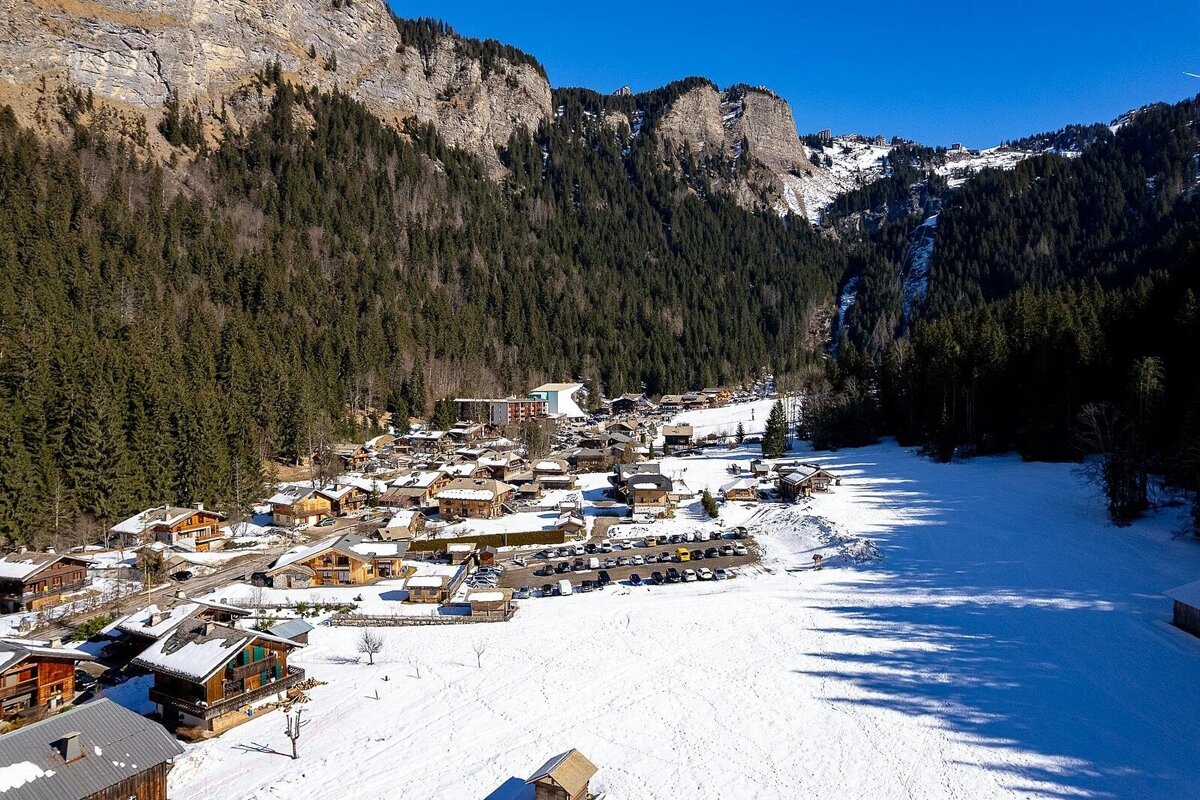 Aerial shot of a charming snowy mountain village with wooden chalets, framed by dense forests and rugged cliffs under a clear blue sky.