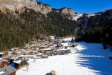 Aerial shot of a charming snowy mountain village with wooden chalets, framed by dense forests and rugged cliffs under a clear blue sky.