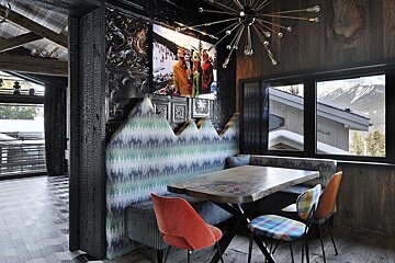 A chic, eclectic mountain dining nook featuring a unique mountain-shaped bench, rustic wood, colorful seating, and a stunning snowy peak view.