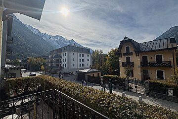 A sunlit view of a mountain town with snow-capped peaks, buildings, and a street, framed by a balcony railing and hedges.