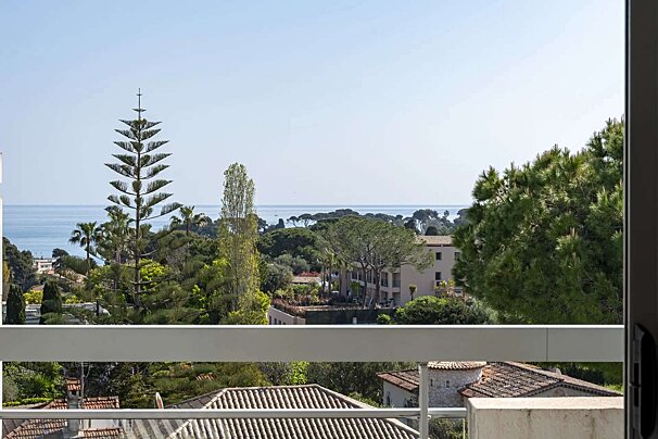 A balcony view reveals a serene coastline with a distinctive tall pine tree, lush greenery, and scattered red-roofed buildings under a clear sky.