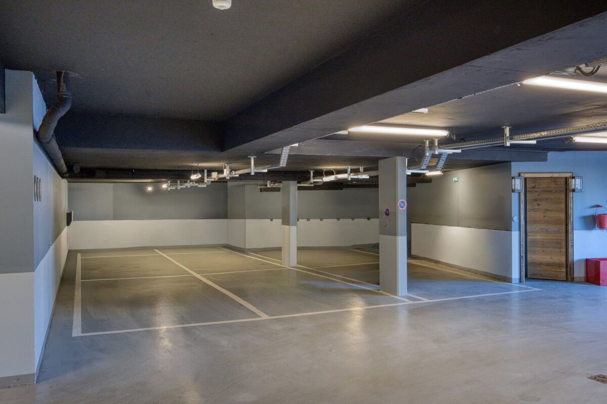 An empty underground parking garage with grey walls, white parking lines, support pillars, and overhead lighting.