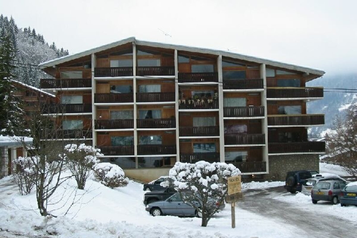 A multi-story wooden building with balconies is covered in snow, surrounded by snow-dusted trees and parked cars in a winter mountain scene.