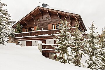 A rustic, multi-level wooden chalet with carved details, heavily covered in fresh snow, surrounded by frosted pine trees on a winter day.